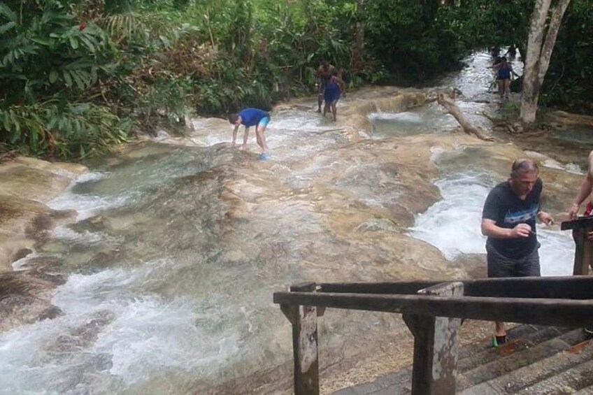 A river with people standing on rocks, a wooden bridge, and lush greenery. 4