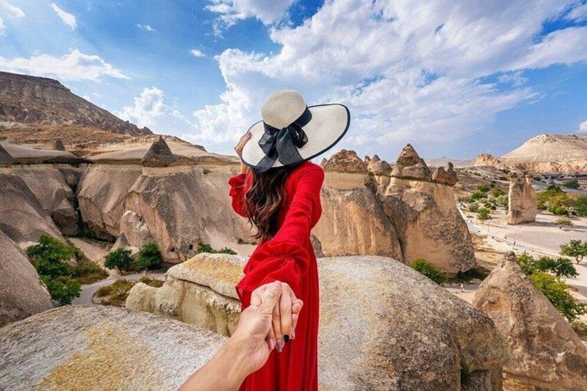 Eine Person in einem roten Kleid hält die Hand einer anderen Person, während sie in einer felsigen Landschaft mit großen Felsen und kargen Pflanzen steht. 3