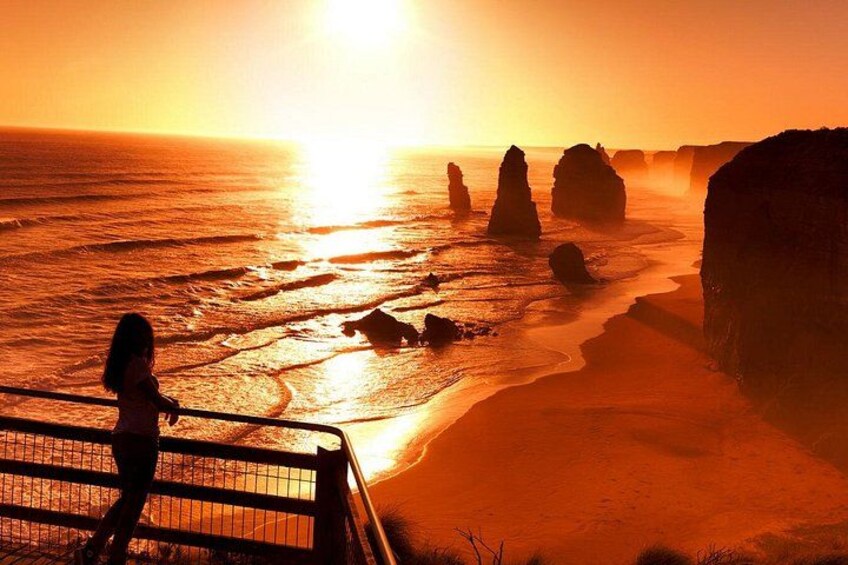 A sunset over a beach with rock formations and a person standing on a railing. 1