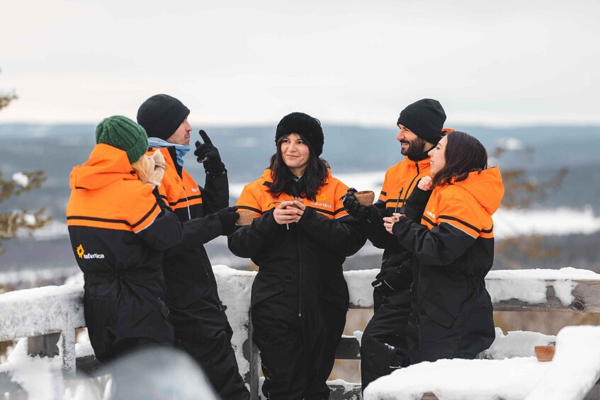 Four people in winter clothing standing on a snow-covered ledge with a mountainous landscape in the background. 5