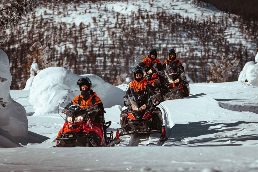 Four people on snowmobiles in a snowy landscape with a forested hill in the background. 2