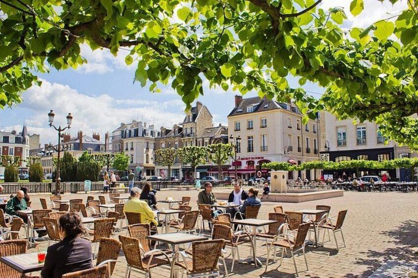 Un café al aire libre soleado con mesas y sillas de mimbre, rodeado de edificios y árboles. 2
