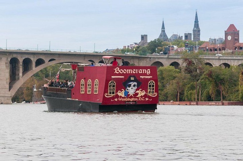 Un barco rojo con la leyenda 'Booerang' escrita, un puente al fondo y un paisaje urbano con edificios y árboles. 1