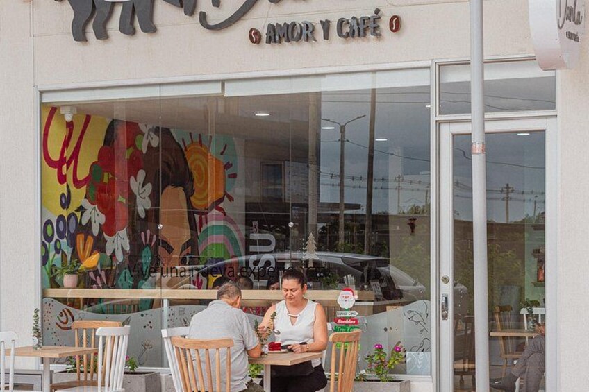 Een café met een kleurrijke muurschildering op de etalage, terras en een geparkeerde auto op de achtergrond. 2