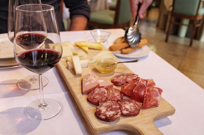 A table with a wooden board of various cheeses and cured meats, a glass of red wine, and a small bowl of honey. 1