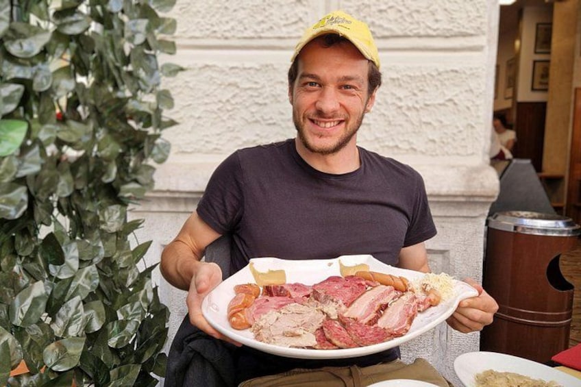 Une personne tenant une assiette remplie de différentes sortes de viande, assise à l’extérieur, avec un bâtiment et de la végétation en arrière-plan. 2