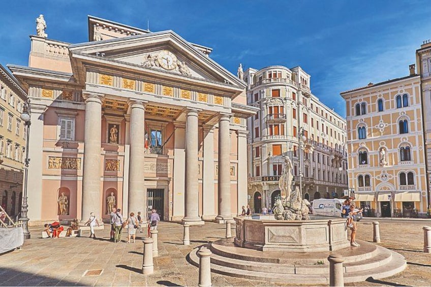 Una plaza histórica con arquitectura clásica, incluyendo un edificio destacado con columnas y una estatua en el centro. 1