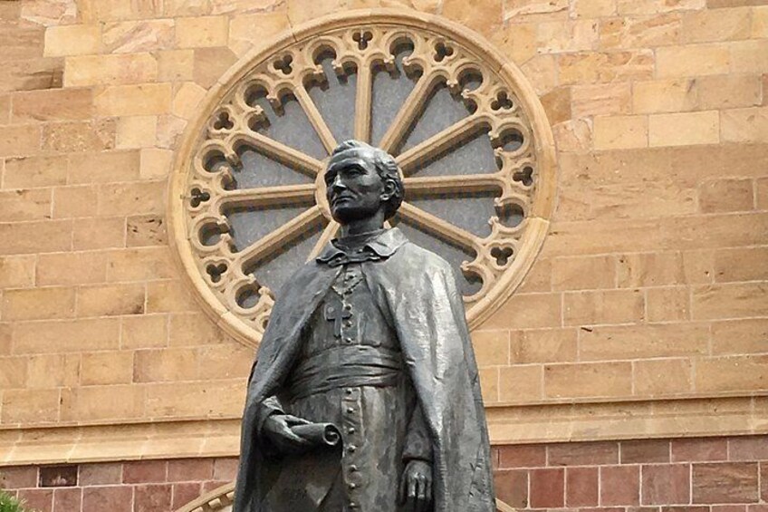 Una estatua de una persona vestida con una túnica y una cruz, de pie frente a un edificio con un ventanal circular grande. 2