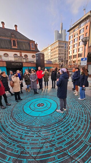 A group of people standing in a circular pattern on a patterned pavement with a building labeled 'MOMEM' in the background. 5