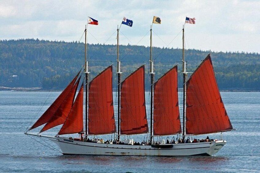 A sailboat with multiple sails, flags, and passengers on a body of water with a forested shoreline in the background. 1