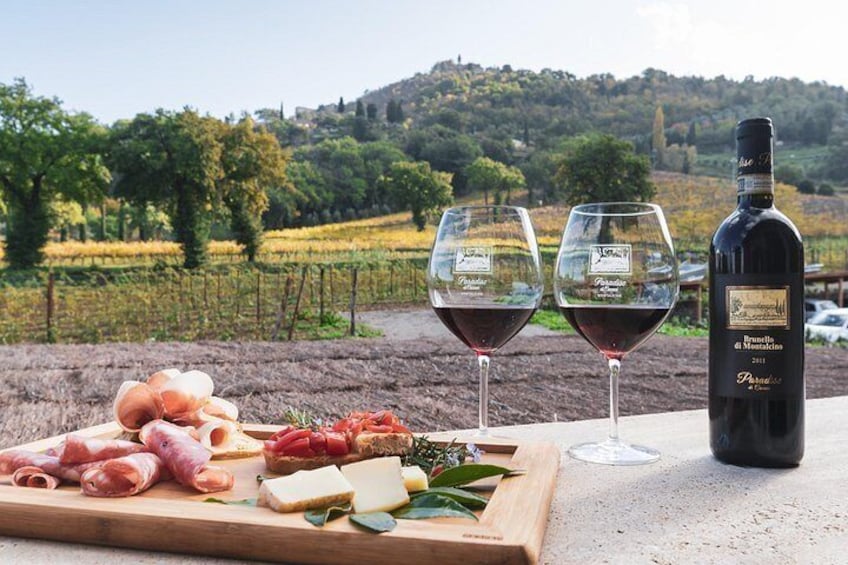 Une bouteille de vin, deux verres de vin rouge et une planche de charcuterie avec des viandes et des fromages, le tout posé sur une table avec un vignoble en arrière-plan. 2