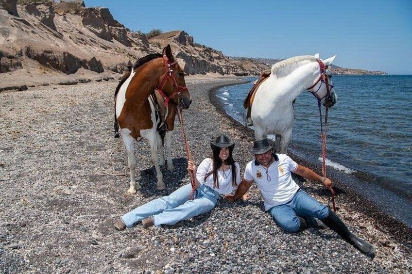 Twee personen zitten op een kiezelstrand, met paarden in de buurt. 2