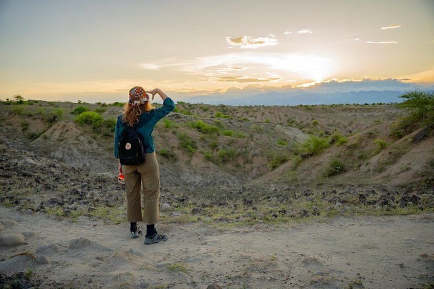 A person standing on a dirt path with a backpack, looking at a sunset over a hilly landscape. 2