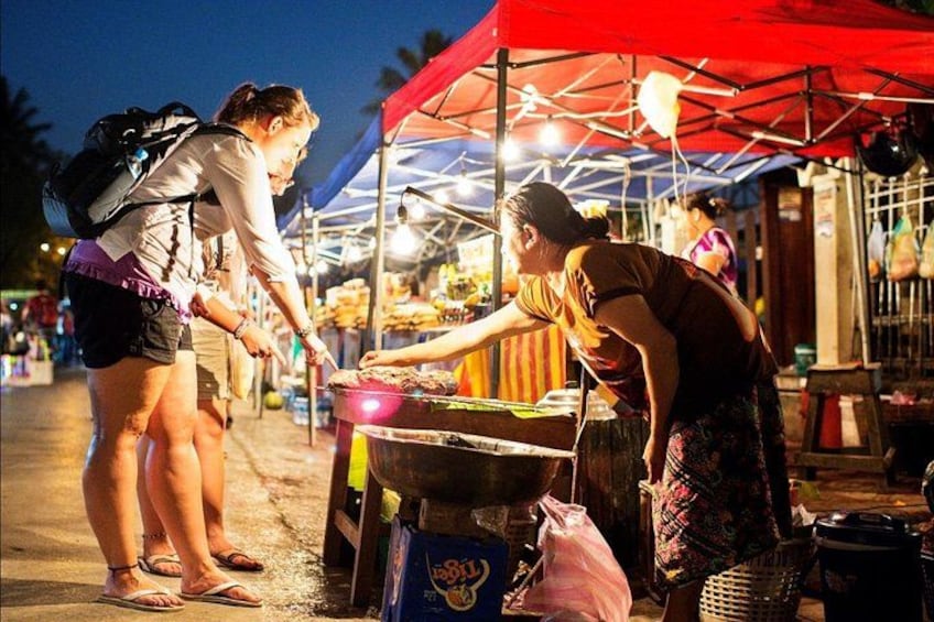Un venditore di cibo di strada cucina a un banco coperto da una tenda rossa, un cliente compra cibo, e un'atmosfera da mercato. 4