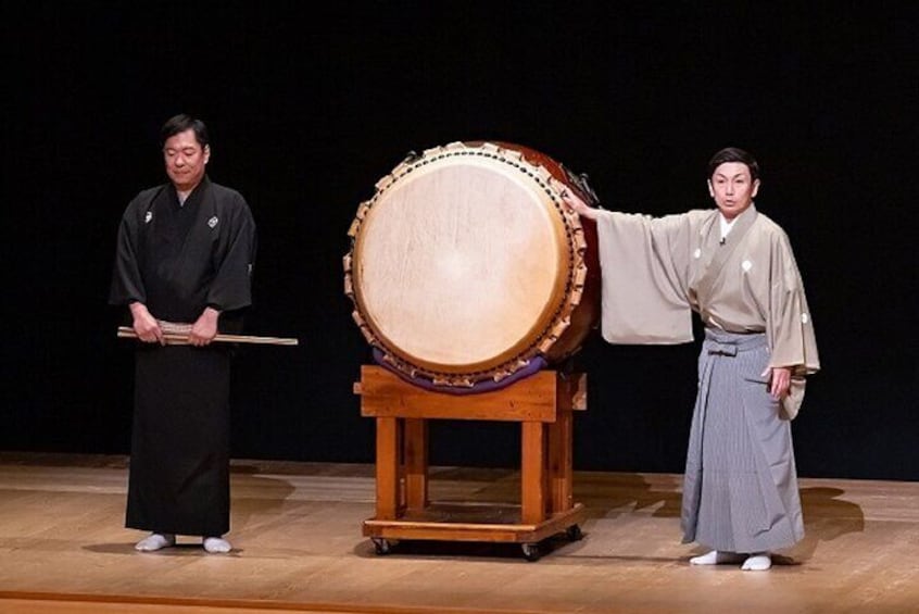 Two individuals in traditional attire, one playing a drum and the other gesturing towards a large drum. 2