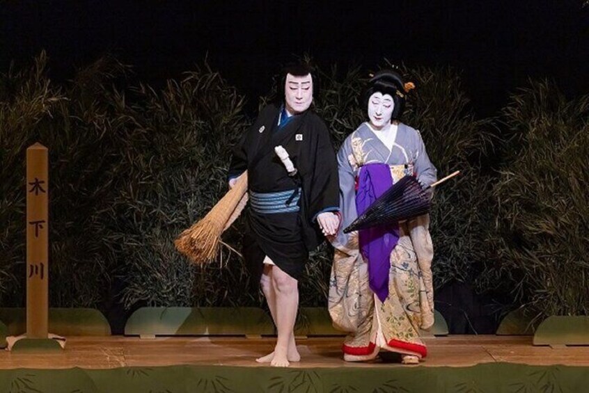 Two performers in traditional Japanese attire, one with a fan and the other with a broom, on a stage with a backdrop of bamboo. 3