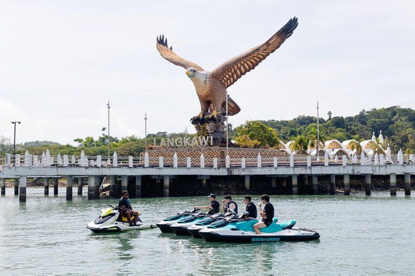 A large bird sculpture on a bridge, people on watercraft, and a sign with 'LANGKAW'. 1
