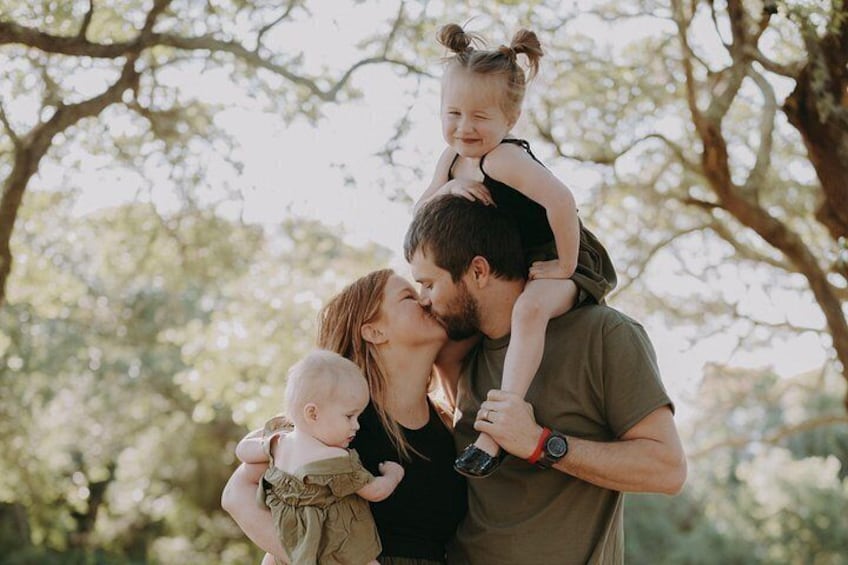 A family outdoors with a man holding two children, one on each shoulder, in a natural setting with trees. 5