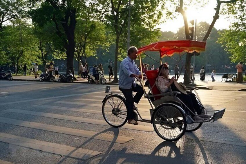 A person pushing a rickshaw with a passenger, a park with trees and people in the background. 4