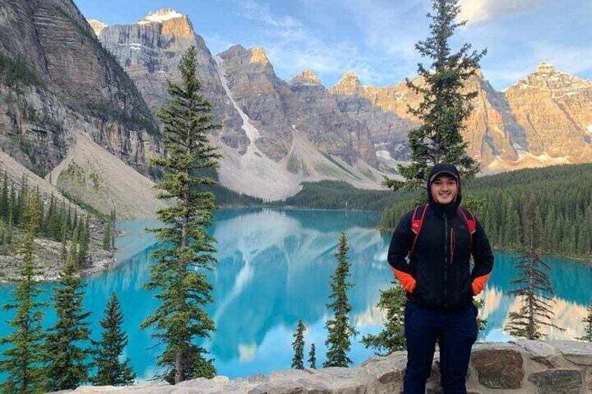 A person standing in front of a mountain lake with clear turquoise water, surrounded by tall trees and rocky terrain. 1