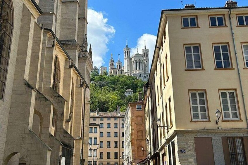 A narrow street with buildings on either side, a church with two towers in the background, and a hill with greenery. 5