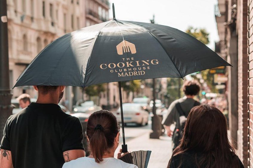 Eine Gruppe von Menschen geht auf einer Stadtstraße, wobei eine schwarze Tasche mit dem Aufdruck 'The Cooking Clubhouse Madrid' zu sehen ist. 1