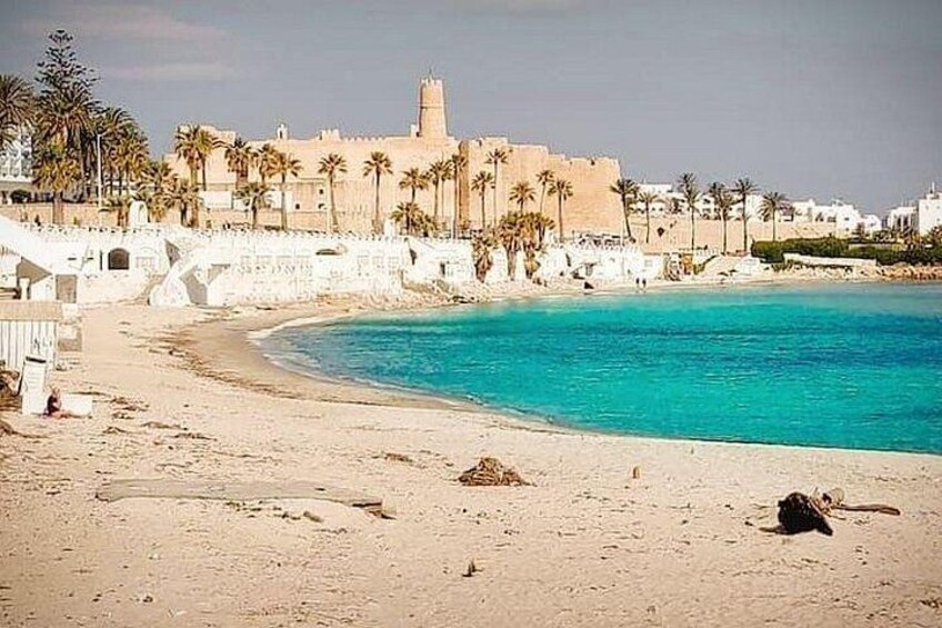 A sandy beach with clear turquoise water, palm trees, and a historic building with a tower in the background. 3
