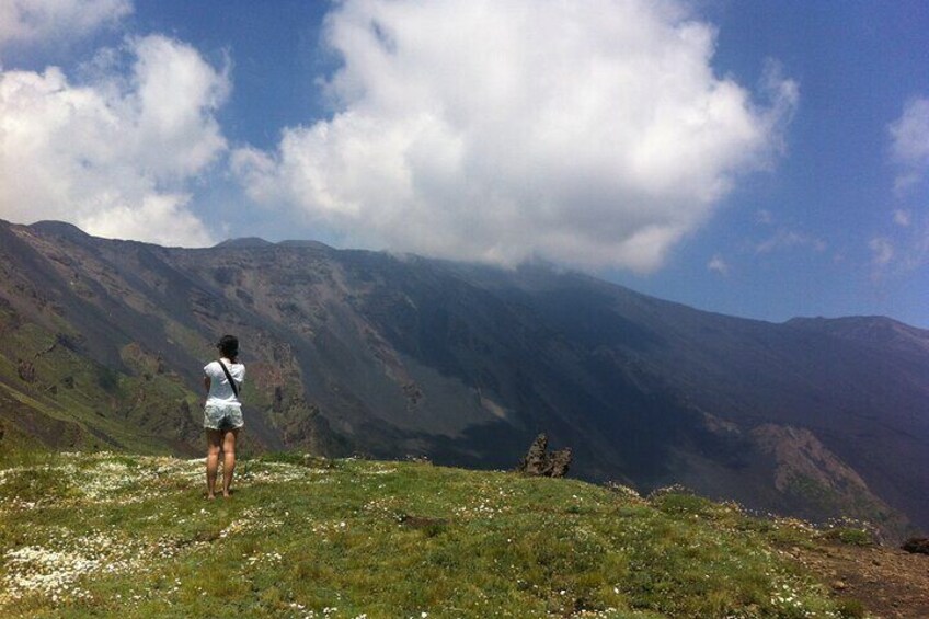 Une personne se tient sur une colline herbeuse, d’où elle domine un paysage montagneux, avec des nuages dans le ciel. 2