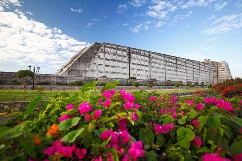 Un edificio grande con techo inclinado, rodeado por un jardín cuidado con flores rosadas. 2