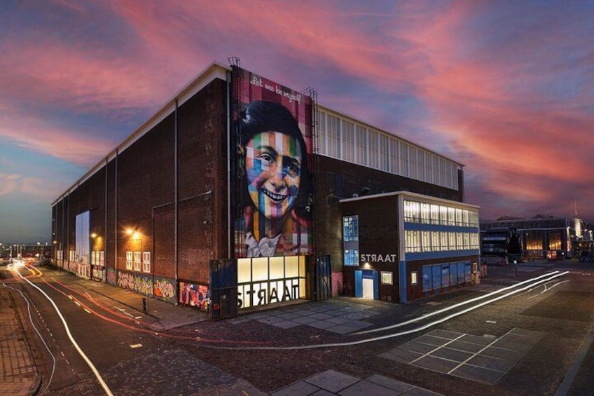 Un bâtiment orné d’une grande fresque murale, une rue illuminée par des traînées de lumière et un ciel au coucher du soleil. 5