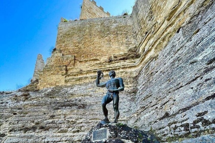 Una estatua de una persona sobre un afloramiento rocoso con formaciones rocosas estratificadas al fondo. 3