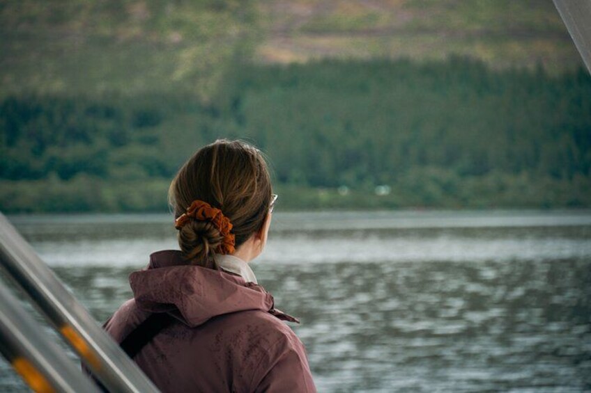 A person in a pink jacket with a flower in their hair, looking out over a body of water with a forested hillside in the background. 3