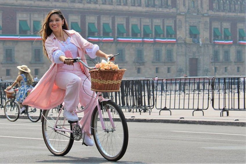 A person riding a bicycle with a basket on a road, with buildings in the background. 5