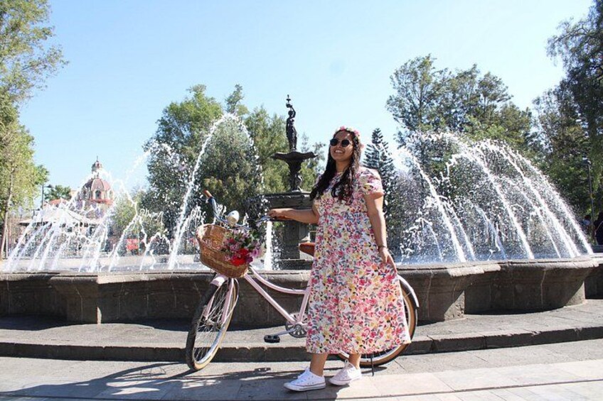 A woman in a floral dress standing next to a fountain with water jets, a bicycle with a basket, and a statue in the background. 2