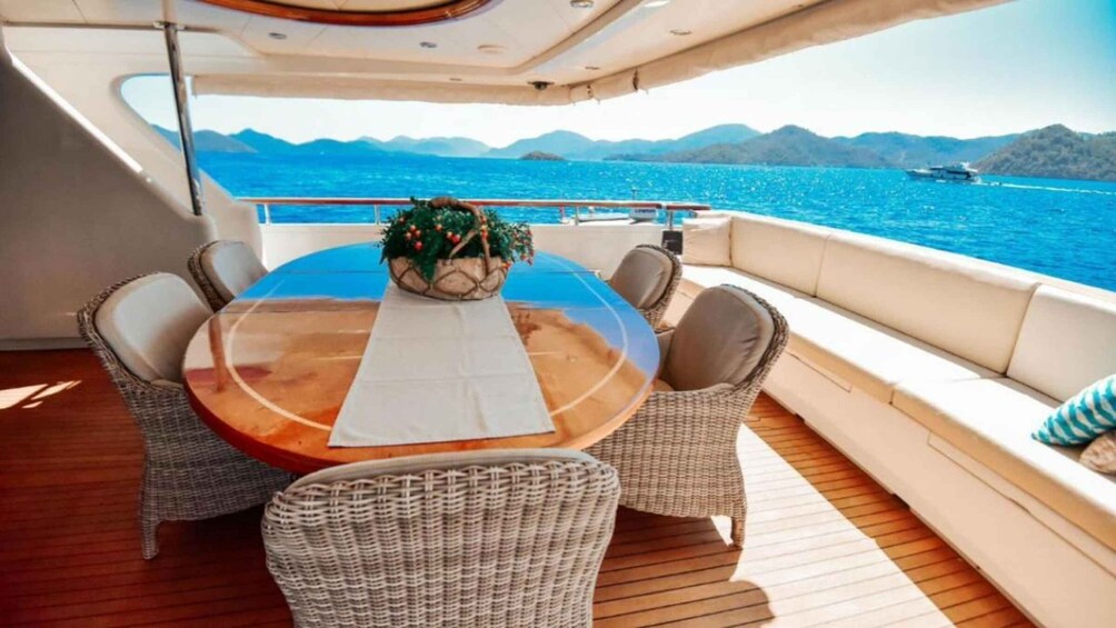 A boat deck with a round table, wicker chairs, a potted plant, and a view of the sea and mountains. 2