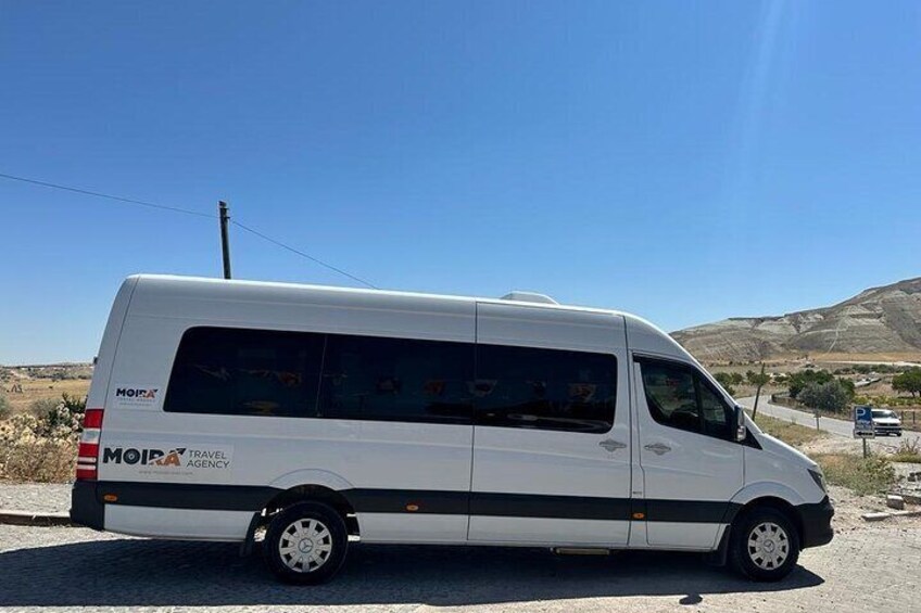 A white Moira Travel Agency van parked on a road with a clear blue sky and distant hills in the background. 3