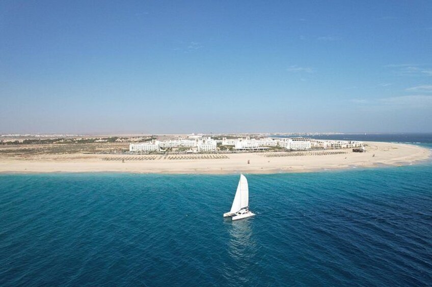 A sailboat on a turquoise sea with a beach and buildings in the background. 3