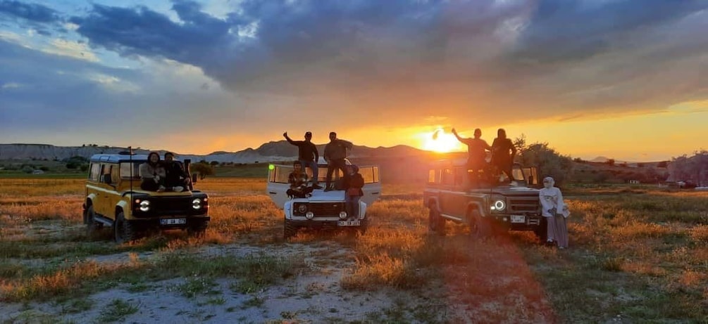Three off-road vehicles parked in a field with people standing on them, and the sun setting in the background. 1