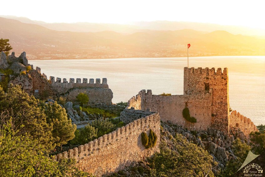 A historic stone fortress with a flag on top, overlooking a body of water with mountains in the background. 4