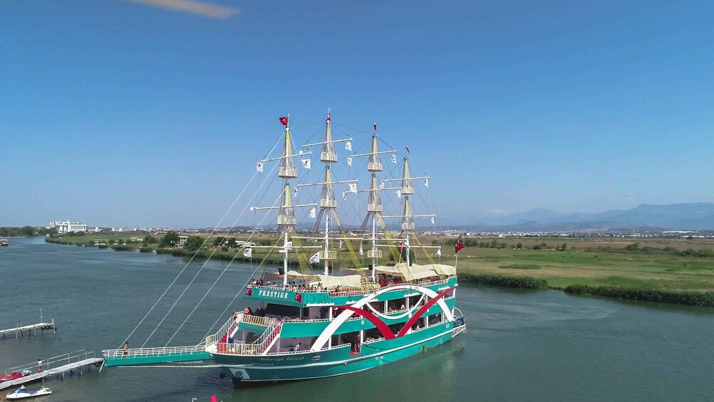 A large, multi-decked sailing ship with multiple masts and flags, docked at a marina with a clear blue sky and distant mountains in the background. 1
