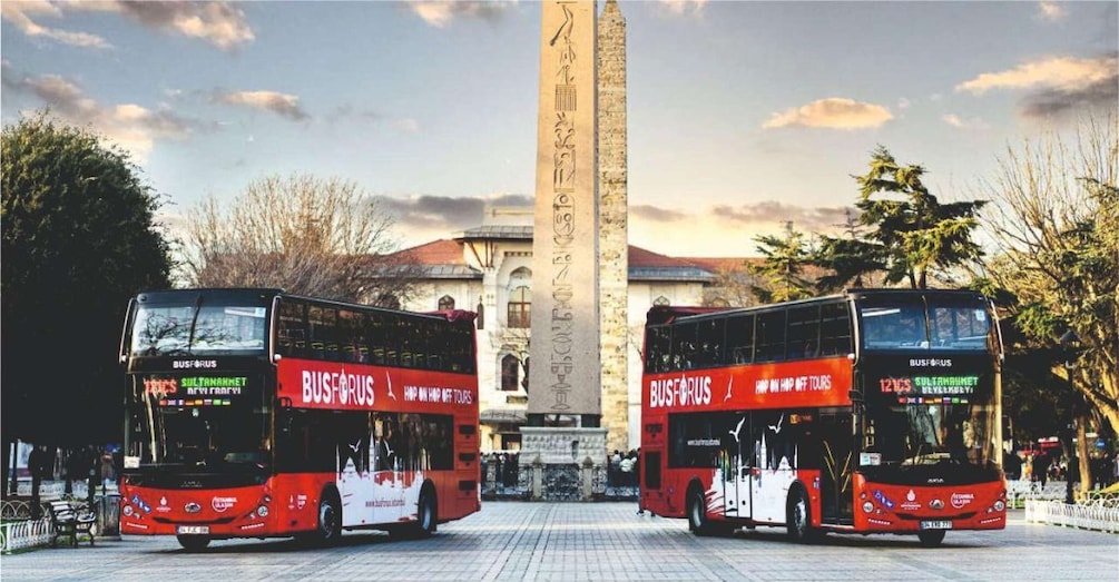 Two red double-decker buses parked in front of an obelisk with hieroglyphs. 2
