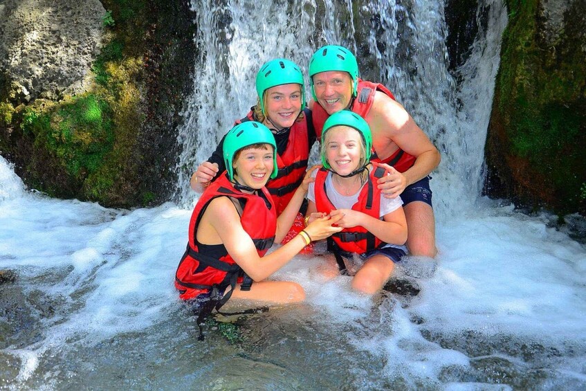 A group of people in red life jackets and green helmets sitting in a stream with a waterfall in the background. 3