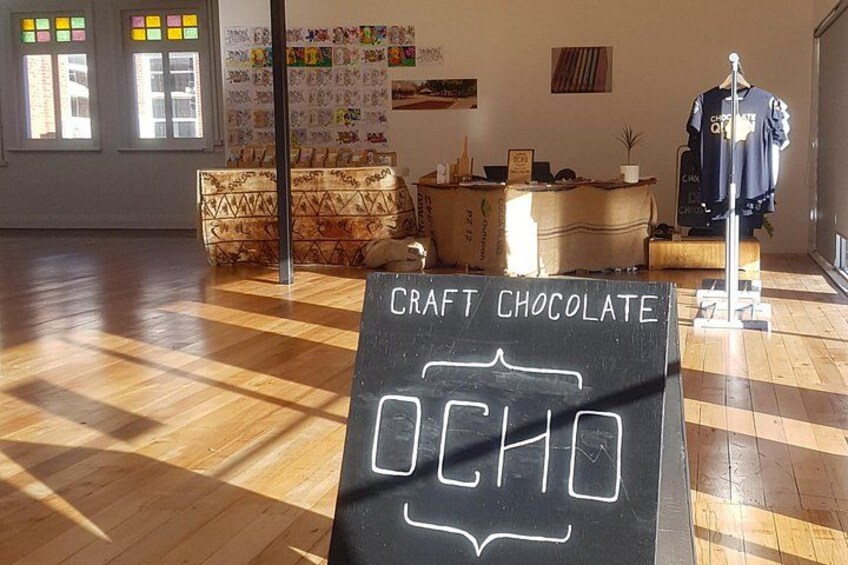 A room with a chalkboard sign reading 'CRAFT CHOCOLATE OCHO,' wooden flooring, a mannequin, and a wall with colorful artwork. 3