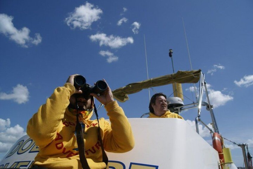 Una persona tomando una fotografía con una cámara, otra persona de pie junto a un velero blanco grande, y un cielo azul despejado con nubes. 3