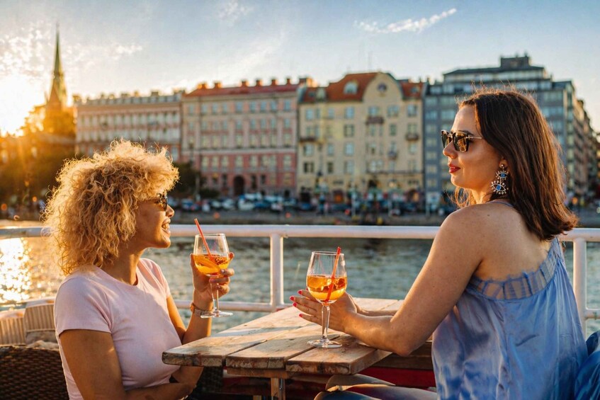 Två personer sitter vid ett träbord vid vattnet och håller i varsin drink. 3
