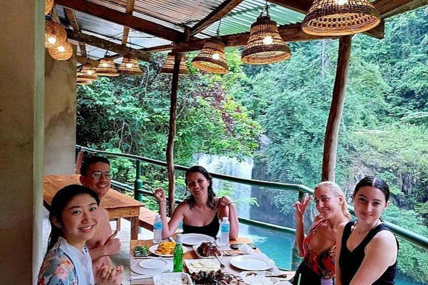 Een groep mensen dineert aan een tafel met uitzicht op een weelderig bos en een waterval. 2