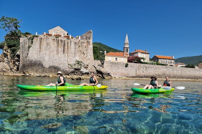 Three people kayaking on clear water with a historic stone wall and buildings in the background. 1