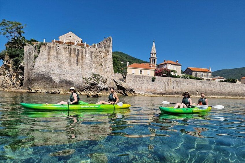 Three people kayaking on a clear lake with a historic stone wall and a church in the background. 1