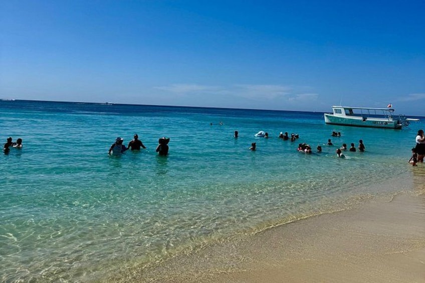 A beach with clear turquoise water, people swimming, and a boat in the distance. 4