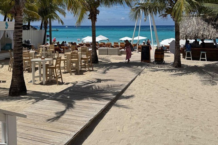 A beach with wooden tables and chairs, palm trees, and umbrellas. 2